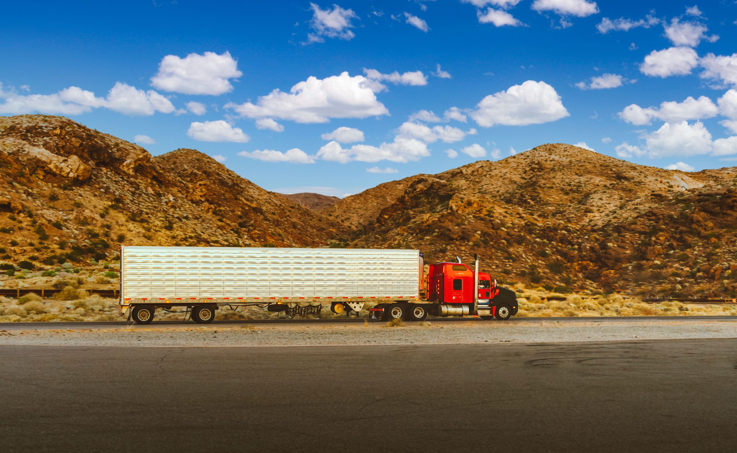 Red semi-truck driving through a desert highway with mountain views, representing a long-haul trucking route across America